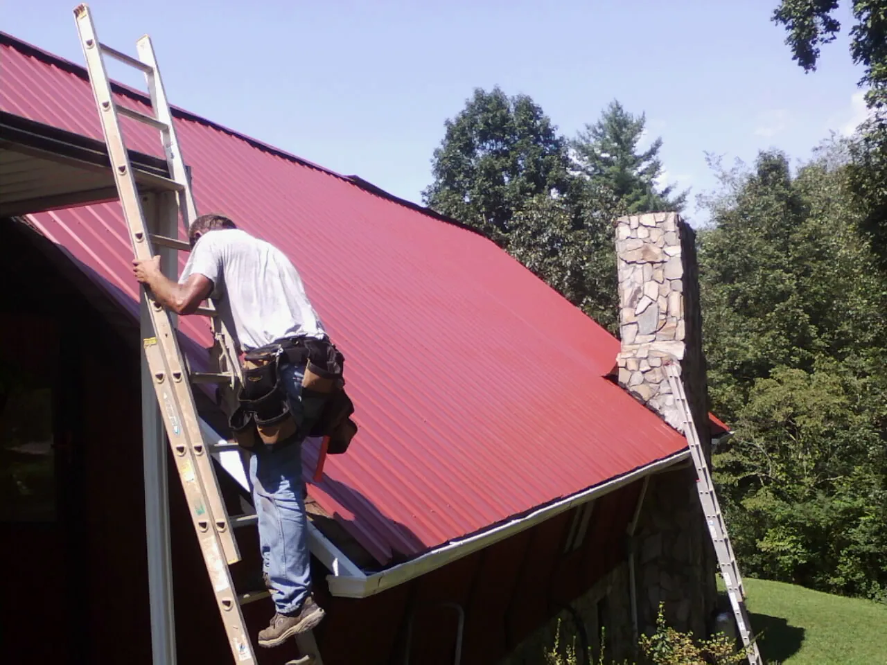 Roofer on a ladder beside a red metal roof and stone chimney — Soffit & Fascia Repair by Roofing Brothers in Dewar
